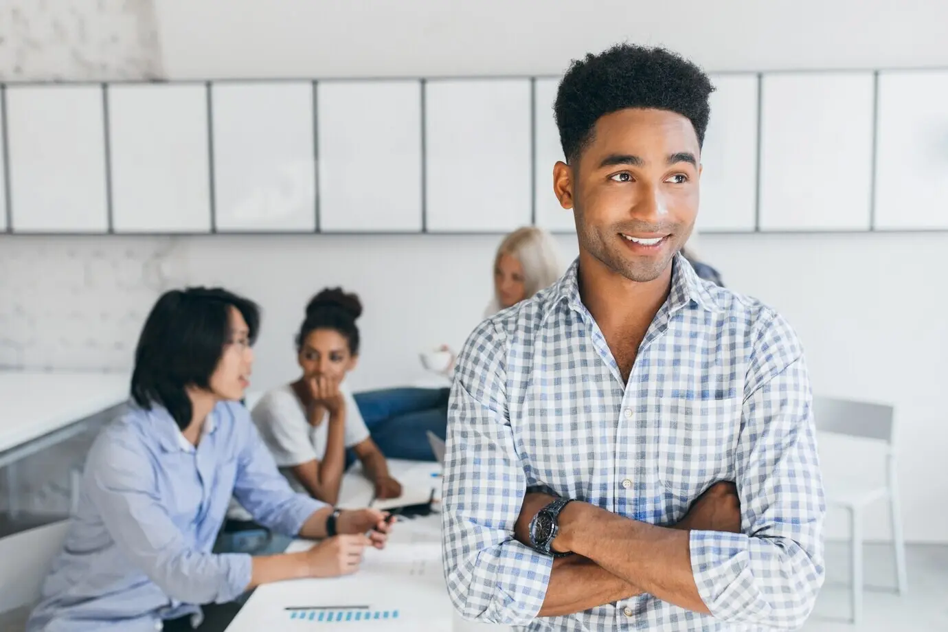 Gutaussehender schwarzer junger Mann mit Armbanduhr, der den Blick abwendet, während seine Kollegen neue Ideen diskutieren. Innenporträt internationaler IT-Spezialisten mit einem afrikanischen Mann.